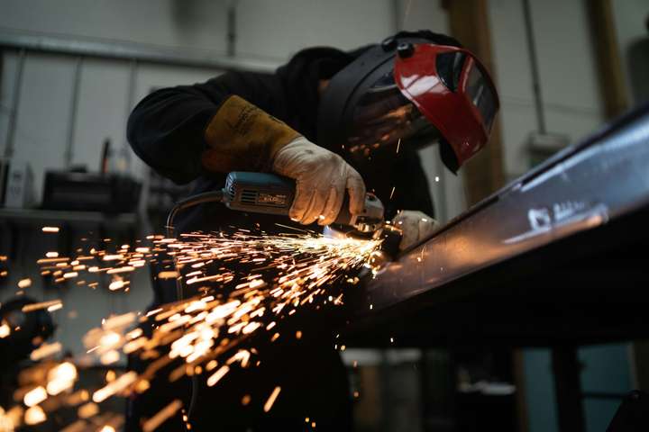 A metalworker in a face shield grinding a steel beam, sparks flying