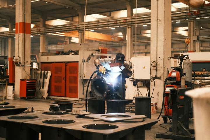A masked welder joining a workpiece at a station in a large metal fabrication hall